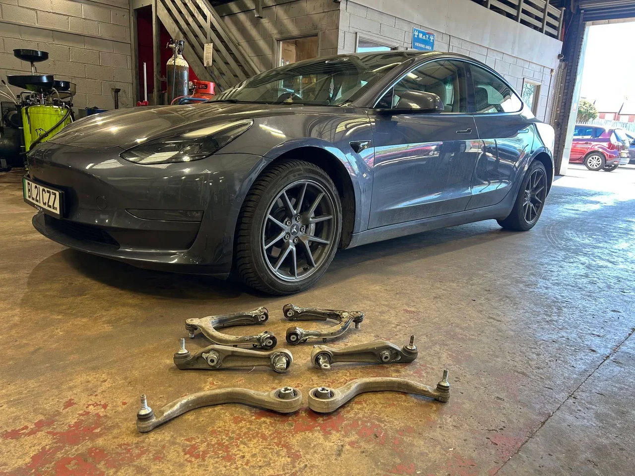 Автомобиль Tesla Model 3 в мастерской KMC Electric; снятые детали подвески разложены перед машиной.