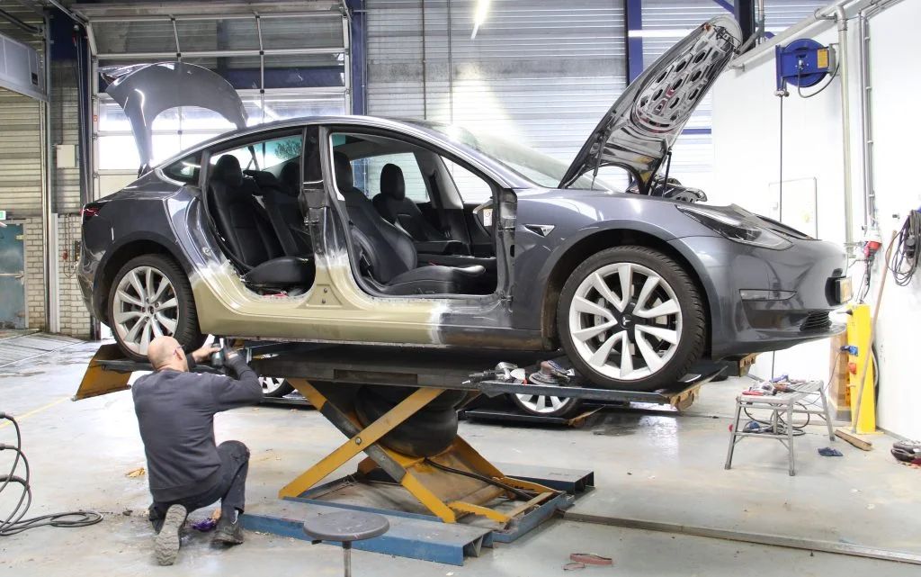 Tesla с открытыми дверями в кузовной мастерской Metropool; мастер работает у автомобиля на подъёмнике.