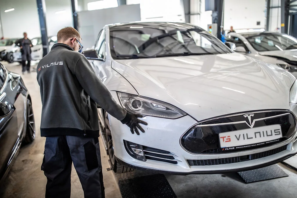 Сотрудник TS Vilnius работает у Tesla внутри мастерской; логотип сервиса виден на куртке и номере автомобиля.