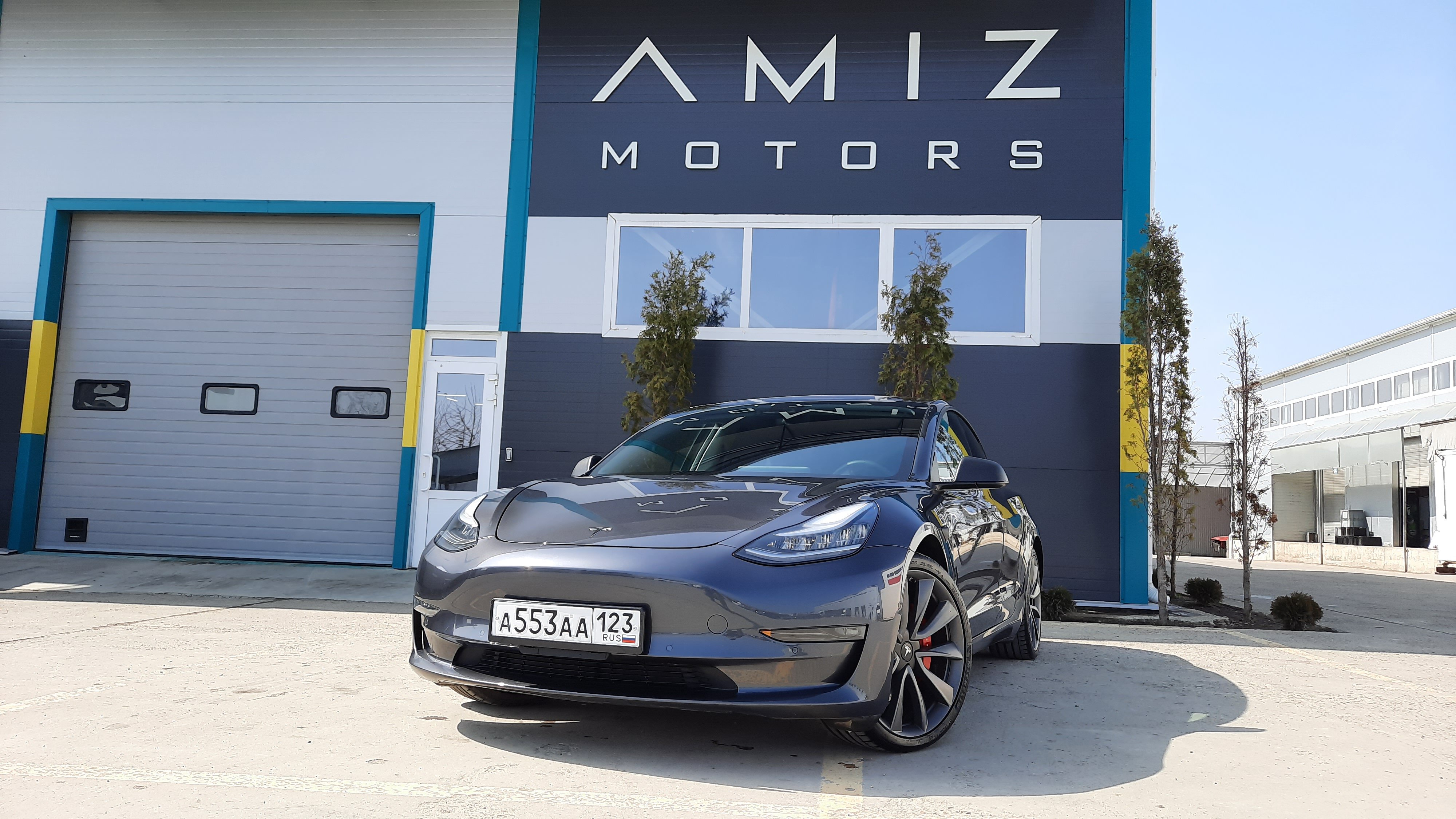 Фасад с вывеской AMIZ MOTORS и припаркованной перед ним Tesla Model 3.