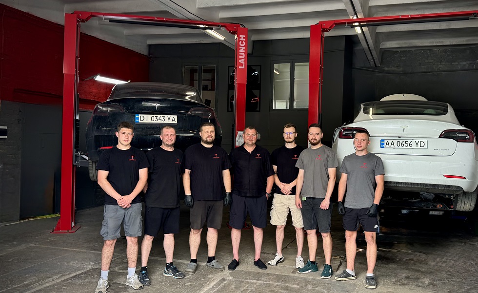 Сотрудники позируют в мастерской рядом с двумя автомобилями Tesla на подъёмниках.