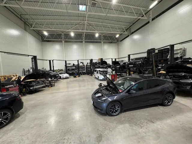EV Workshop darba zona Rīgā: vairāki Tesla automobiļi uz pacēlājiem un Tesla Model 3 ar atvērtu motora pārsegu. Foto uzņemts 2025. gada septembrī.