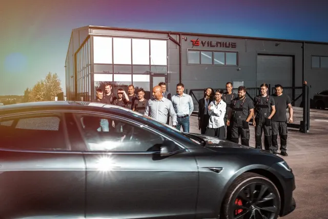 TS Vilnius komanda prie aptarnavimo pastato su aiškiai matomu logotipu; priekyje Tesla automobilis.