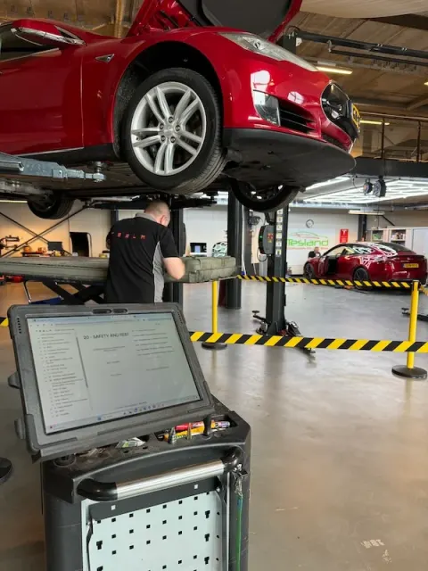 Een rode Tesla op een hefbrug bij Tesland, met een monteur en diagnoseapparatuur in de buurt.