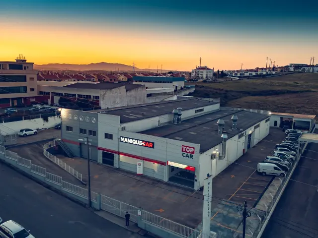 Vista aérea da oficina de carroçaria Maniquecar e do parque de estacionamento; o letreiro é visível na fachada.